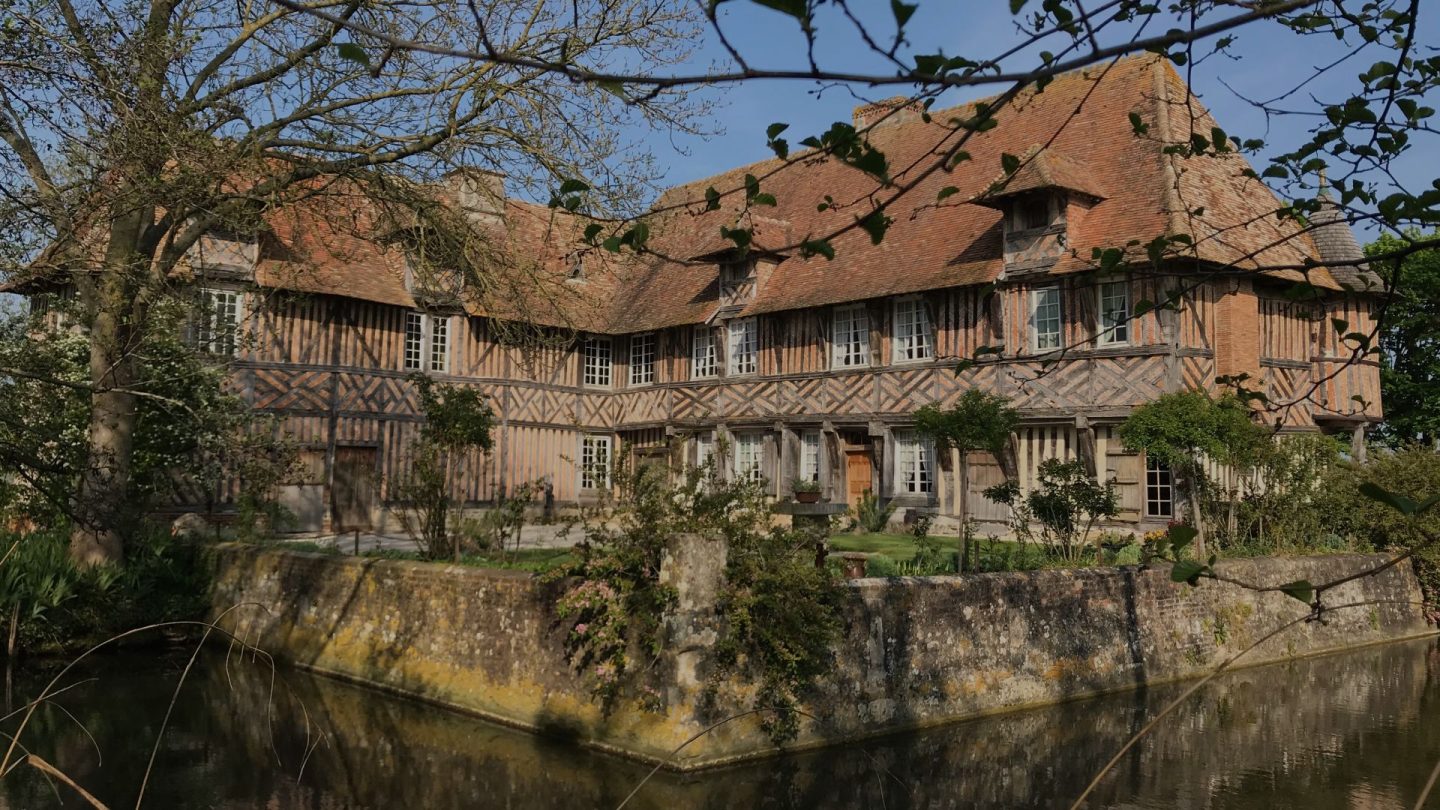 Le manoir de Coupesarte vu depuis les douves, ses deux ailes à pans de bois sur leur plateforme.